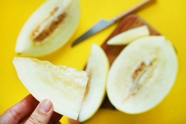 Ahşap bir tahtanın üzerinde dilimlenmiş sarı kavunun arka planında bir kadının elinde bir parça kavun.