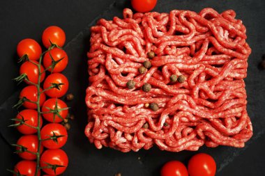 Koyu arkaplanda kiraz domatesleri ve baharatların yanında siyah bir tepside taze kıyma.