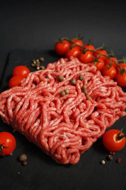Koyu arkaplanda kiraz domatesleri ve baharatların yanında siyah bir tepside taze kıyma.