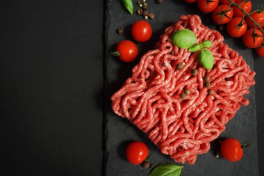 Koyu arkaplanda kiraz domatesleri, fesleğen ve baharatların yanında siyah bir tepside taze kıyma.