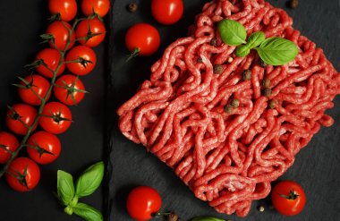 Koyu arkaplanda kiraz domatesleri, fesleğen ve baharatların yanında siyah bir tepside taze kıyma.