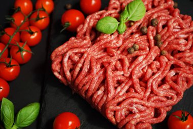 Koyu arkaplanda kiraz domatesleri, fesleğen ve baharatların yanında siyah bir tepside taze kıyma.