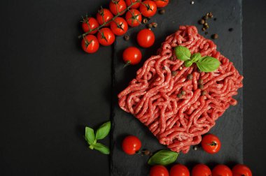 Koyu arkaplanda kiraz domatesleri, fesleğen ve baharatların yanında siyah bir tepside taze kıyma.