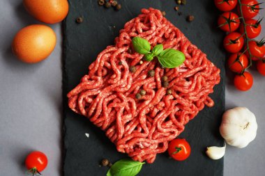 Siyah bir tepside taze kıyma tepsisi üzerinde, gri arka planda kiraz domatesleri, fesleğen, yumurta ve baharatların yanında.