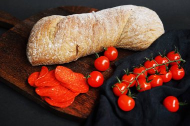 Tahtada taze ekmek, kiraz domateslerinin yanında ve arka planda salam dilimleri.