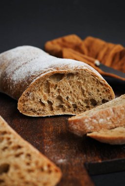 Koyu arkaplanda keten peçetesi olan bir bıçağın yanındaki tahta tahtada taze kesilmiş ekmek.