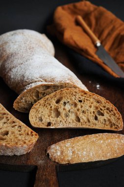 Koyu arkaplanda keten peçetesi olan bir bıçağın yanındaki tahta tahtada taze kesilmiş ekmek.