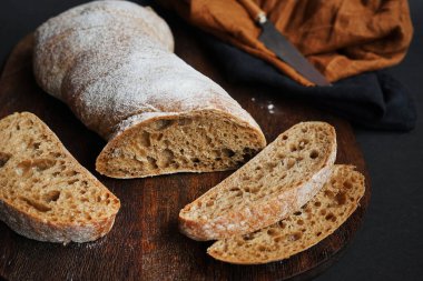 Koyu arkaplanda keten peçetesi olan bir bıçağın yanındaki tahta tahtada taze kesilmiş ekmek.