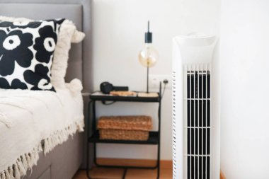 Beyaz kule vantilatörü, yatak ve komodinin yanında, içinde lamba olan modern, parlak bir iç mekan.