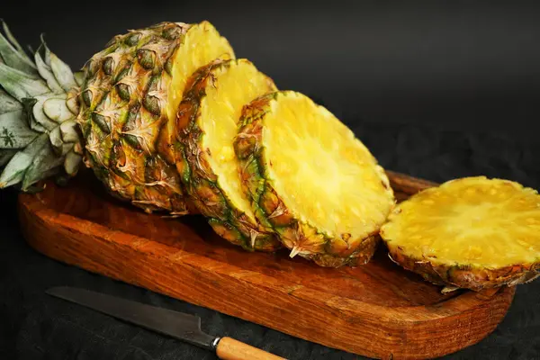 Olgun ananas, tahta tepside dilimlenmiş, koyu arkaplanda bir bıçağın yanında.