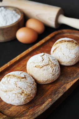 Yumurtaların yanındaki ahşap tepside küçük taze ekmekler, unlu ahşap bir kase ve merdane.