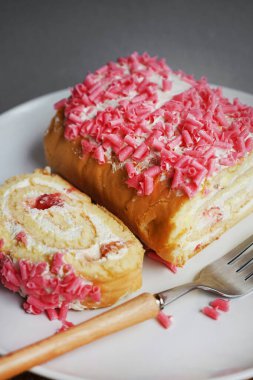 Kremalı ve pembe parçalı sünger rulosu beyaz bir tabakta, gri arka planda bir çatalın yanında.