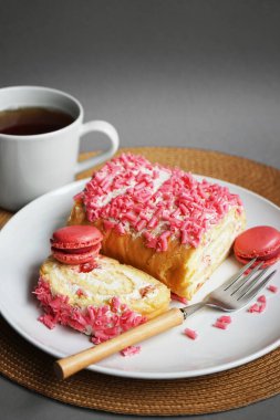 Kremalı sünger rulosu, pembe serpinti ve ahudududulu kurabiye beyaz bir tabakta, çatal ve fincanın yanında gri bir arka planda.