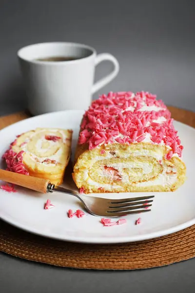 stock image Sponge roll with cream and pink sprinkles on a white plate next to a fork and cup on a gray background