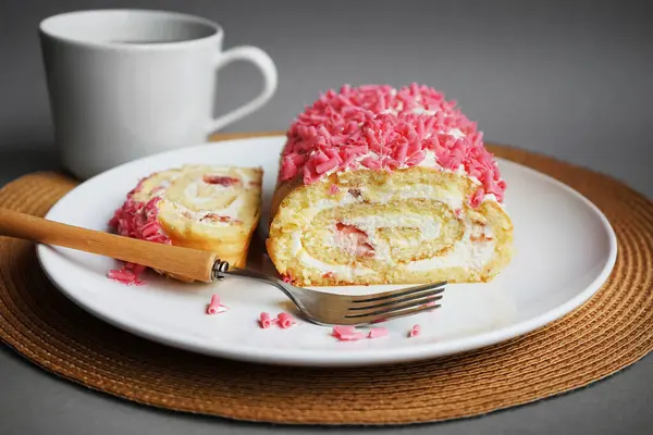 stock image Sponge roll with cream and pink sprinkles on a white plate next to a fork and cup on a gray background