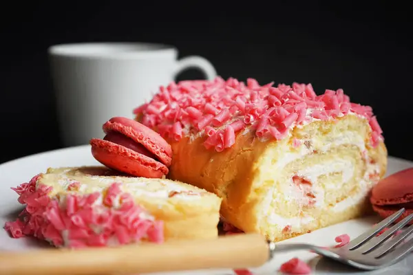 Stock image Sponge roll with cream, pink sprinkles and raspberry macaroons on a white plate next to a fork and cup on a dark background