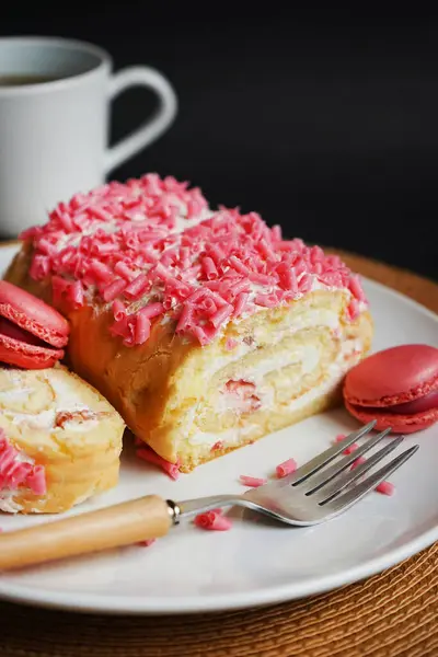 stock image Sponge roll with cream, pink sprinkles and raspberry macaroons on a white plate next to a fork and cup on a dark background