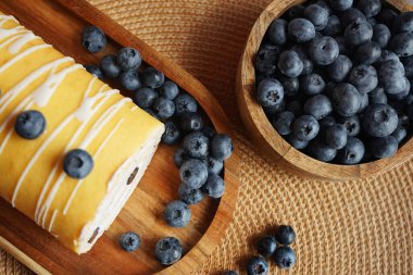 Krema ve yaban mersinli sünger rulosu ahşap tepside, bej bir peçetenin yanında gri bir arka planda.