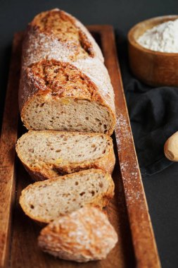 Tahta tepside taze dilimlenmiş ekmek, bir kase un ve koyu arkaplanda tahta bir oklavanın yanında.