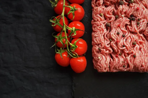 stock image Minced meat with black and red peppers on a black slate tray next to a branch of tomatoes on a dark background