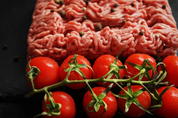 Stock image Minced meat with black and red peppers on a black slate tray next to a branch of tomatoes on a dark background