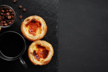 Siyah bir fincan kahvenin yanında yumurtalı tartlar ve koyu bir arka planda dağınık kahve çekirdekleri.