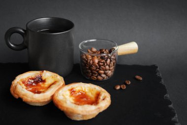 Siyah bir fincan kahvenin yanında yumurtalı tartlar ve koyu bir arka planda dağınık kahve çekirdekleri.