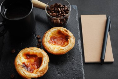 Koyu kahve fincanının yanında yumurtalı tartlar, dağınık kahve çekirdekleri ve koyu arkaplanda bir not defteri.