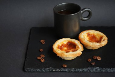 Siyah bir fincan kahvenin yanında yumurtalı tartlar ve koyu bir arka planda dağınık kahve çekirdekleri.