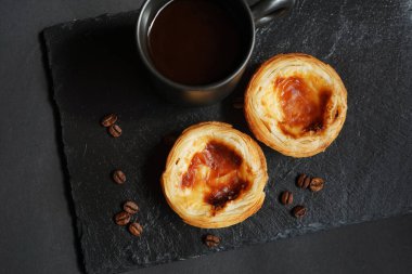 Siyah bir fincan kahvenin yanında yumurtalı tartlar ve koyu bir arka planda dağınık kahve çekirdekleri.