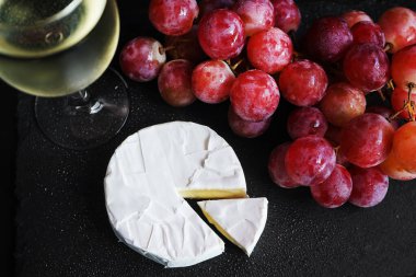 Brie peynirinin yanında bir demet büyük pembe üzüm ve koyu arkaplanda bir bardak beyaz şarap.