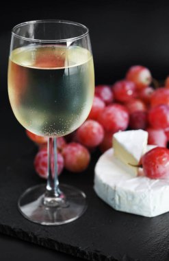 Brie peynirinin yanında bir demet büyük pembe üzüm ve koyu arkaplanda bir bardak beyaz şarap.