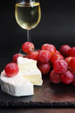 Brie peynirinin yanında bir demet büyük pembe üzüm ve koyu arkaplanda bir bardak beyaz şarap.