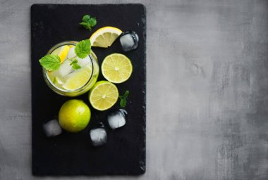 Kola, buz, nane, limon ve kireç dilimli bardak narenciye ve buz küplerini kesmek için gri bir arka planda siyah bir tepside.