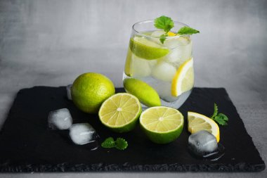 Kola, buz, nane, limon ve kireç dilimli bardak narenciye ve buz küplerini kesmek için gri bir arka planda siyah bir tepside.