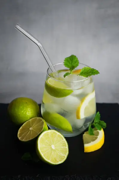 Sodalı, buzlu, naneli, limonlu ve limonlu bardaklar, gri arka plandaki siyah tepside narenciye meyvelerini kesmek için.