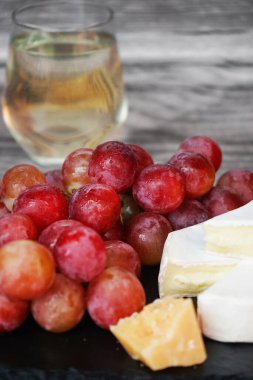 Bir bardak beyaz şarabın yanında büyük pembe üzümler Brie peyniri ve parmesan peyniri gri ahşap arka planda siyah bir tepside.