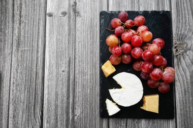 Brie ve Parmesan peynirinin yanında büyük pembe üzümler var. Gri ahşap bir arka planda siyah tepside.