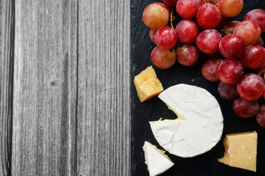 Brie ve Parmesan peynirinin yanında büyük pembe üzümler var. Gri ahşap bir arka planda siyah tepside.