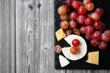 Brie ve Parmesan peynirinin yanında büyük pembe üzümler var. Gri ahşap bir arka planda siyah tepside.