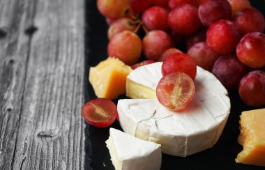 Brie ve Parmesan peynirinin yanında büyük pembe üzümler var. Gri ahşap bir arka planda siyah tepside.