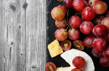 Brie ve Parmesan peynirinin yanında büyük pembe üzümler var. Gri ahşap bir arka planda siyah tepside.