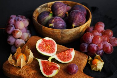 Pembe üzümlerin yanında olgun ve dilimlenmiş incirler ve arka planda tahta bir tahtanın üzerinde Physalis meyveleri.