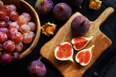 Pembe üzümlerin yanında olgun ve dilimlenmiş incirler ve arka planda tahta bir tahtanın üzerinde Physalis meyveleri.