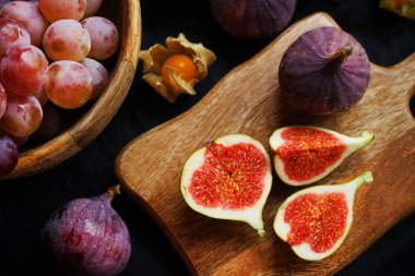 Pembe üzümlerin yanında olgun ve dilimlenmiş incirler ve arka planda tahta bir tahtanın üzerinde Physalis meyveleri.