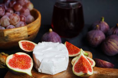 Olgun ve dilimlenmiş incir brie peyniri, Physalis meyvesi, ahşap bir tahtada pembe üzümler ve koyu arkaplanda bir bardak kırmızı şarap.