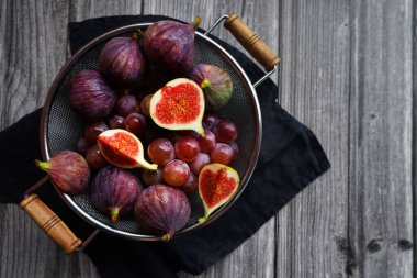 Olgunlaşmış ve dilimlenmiş incirler, ahşap bir arka planda metal bir kevgirde pembe üzümlerin yanında.