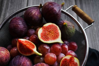 Olgunlaşmış ve dilimlenmiş incirler, ahşap bir arka planda metal bir kevgirde pembe üzümlerin yanında.