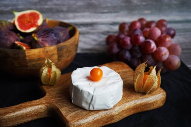 Tahtadaki Brie peynirinin yanında olgun incirler, pembe üzümler ve arka planda Physalis meyveleri.