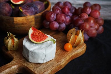 Tahtadaki Brie peynirinin yanında olgun incirler, pembe üzümler ve arka planda Physalis meyveleri.